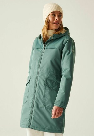 Frau trägt einen türkisfarbenen Wintermantel mit Kapuze und Fronttaschen sowie eine weiße Strickmütze und blickt seitlich vor einem einfarbigen Hintergrund.