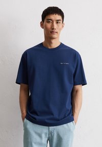 Navyblauwe t-shirt met korte mouwen, gemaakt van zachte katoen, met een ronde hals en een klein wit logo op de borst. Gecombineerd met lichtblauwe jeans.