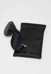 Zwarte oversized zonnebril met donker glas en gouden accenten, rustend op een zwart lederen etui met de opdruk "QUAY." Gladde textuur.