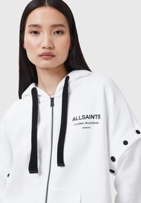 Sudadera blanca con cremallera y cordones negros, bolsillos frontales y detalles de logotipos. Tejido texturizado con un corte relajado y botones decorativos en las mangas.