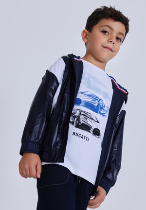 Ragazzo giovane che indossa un giubbotto a vento blu navy e una maglietta bianca con schizzi blu e neri di auto Bugatti, in piedi su uno sfondo chiaro e uniforme.