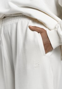Eine dunkelhäutige Hand ruht in der Tasche einer cremefarbenen Jogginghose mit elastischem Bund und dem eingestickten "kamak"-Logo in der Nähe der Tasche.