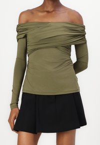 Oliwkowa bluzka typu off-the-shoulder z długimi rękawami, z marszczeniami wokół wcięcia w talii, zestawiona z czarną spódnicą plisowaną.