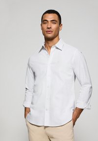Camicia bianca con bottoni, caratterizzata da sottili righe verticali, maniche lunghe arrotolate ai polsini e un piccolo logo sul petto.