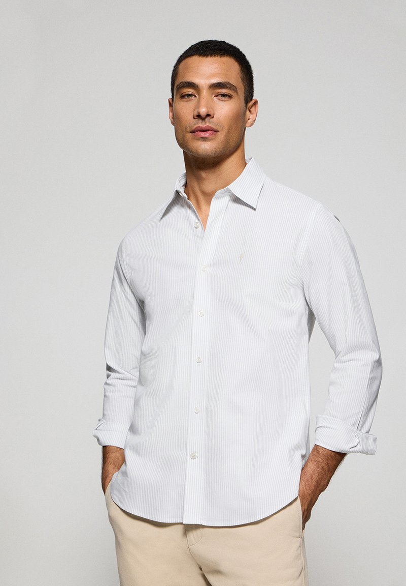 Camicia bianca con bottoni, caratterizzata da sottili righe verticali, maniche lunghe arrotolate ai polsini e un piccolo logo sul petto.