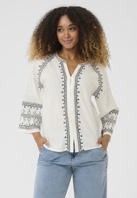 Blusa bianca ampia con motivi ricamati neri lungo le maniche e la patta anteriore; scollatura a V e maniche a tre quarti.
