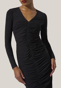 Kvinna klädd i en figurnära svart rynkad långärmad klänning med V-ringning, poserande med händerna nära höfterna mot en neutral bakgrund.