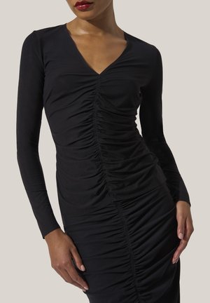 Vrouw in een aansluitende zwarte geplooide lange mouw jurk met V-hals, poseert met haar handen bij haar heupen tegen een effen achtergrond.