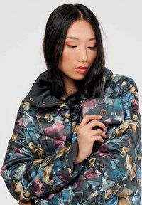 Chaqueta bomber acolchada con un patrón abstracto colorido en azul, rosa y dorado; cuello negro. Sosteniendo una pequeña cartera con cremallera en colores similares.