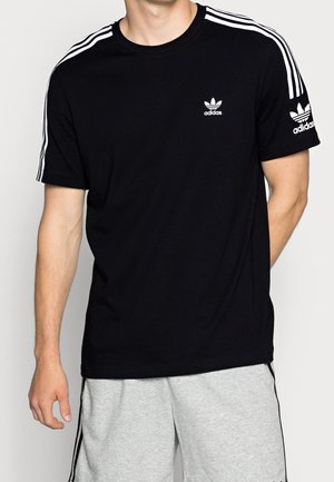 Homme portant un t-shirt Adidas noir avec des bandes blanches sur les épaules et un logo sur la poitrine et la manche, assorti à un short gris clair.