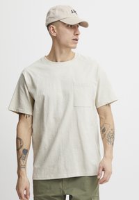 Beige T-shirt med korte ærmer og brystlomme, lavet af blødt stof. Passer godt til olivengrønne bukser. Modellen bærer en beige kasket med sort skrift.