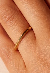Gouden ring met een gestructureerd, gemarmerd oppervlak dat rust op een tan hand, met een glad, cirkelvormig ontwerp en een gepolijste afwerking.
