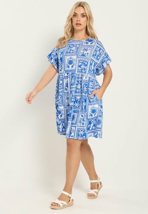 Blondynka w niebiesko-białej sukience o długości do kolan z wzorem, w białych sandałach, stojąca na jednolitym jasnym tle.