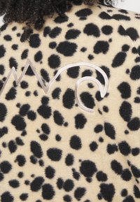 Fleece látka s leopardím potiskem v béžové barvě s černými skvrnami. Obsahuje stříbrnou výšivku tvořící abstraktní tvar vlny na zadní straně.