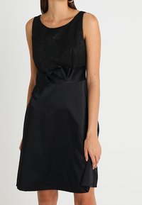 Czarna sukienka bez rękawów z teksturowaną górą i gładką spódnicą, ozdobiona wiązaniem w talii i nieco rozkloszowanym dołem.