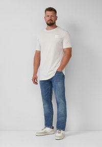 Lichtbeige t-shirt met korte mouwen en een logo op de borst, gecombineerd met blauwe rechttoe-rechtaan jeans en witte sneakers met accentkleuren.