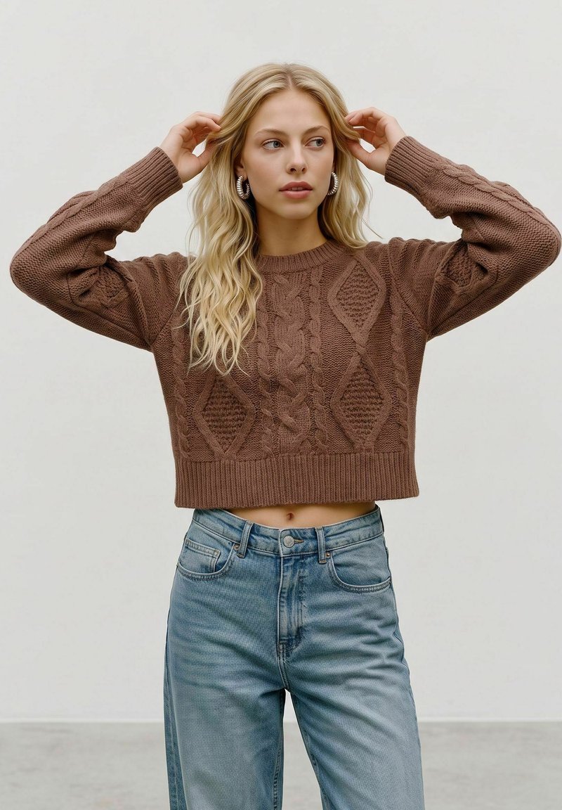 Jeune femme aux longs cheveux blonds porte un pull court marron en maille torsadée, un jean bleu taille haute et des boucles d'oreilles argentées en forme d'anneaux, posant les mains près de la tête.