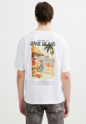 Persona con camiseta blanca con estampado de estilo vintage de una escena de playa, personas, palmeras y texto "Bienvenido a la isla de Oahu."