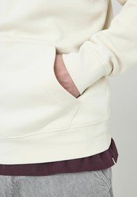 Haut à capuche couleur crème avec une poche avant ; présente un ourlet et des poignets côtelés ; superposé sur une chemise bordeaux et associé à un pantalon gris clair.