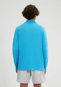 Langärmliges Hemd aus leuchtend blauem Stoff mit klassischem Kragen und lässiger Passform, von hinten gezeigt. Kombiniert mit grauen Shorts.