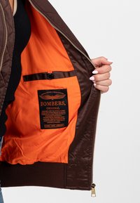 Bruine bomberjack met een gequiltte buitenkant. De binnenkant heeft een helder oranje voering en een zwart etiket met tekst en logo. Ritssluiting.