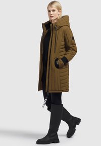 Giacca khaki imbottita con cappuccio, chiusura con zip e tasche laterali; abbinata a leggings neri e stivali neri al ginocchio. Tessuto testurizzato.