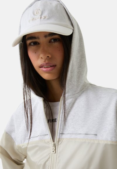 Lichtgrijze zip-up hoodie met een crèmekleurig onderste gedeelte, met een gestreept ontwerp. Het model draagt een witte pet met geborduurd logo en een graphic t-shirt.