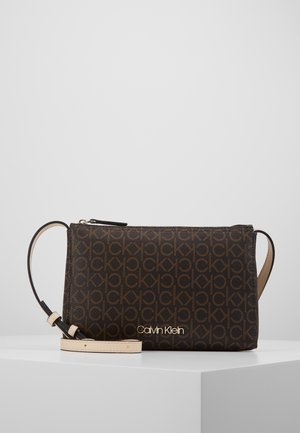 Sac bandoulière marron Calvin Klein avec motif monogramme, logo doré et sangle beige réglable sur un piédestal blanc sur fond gris.