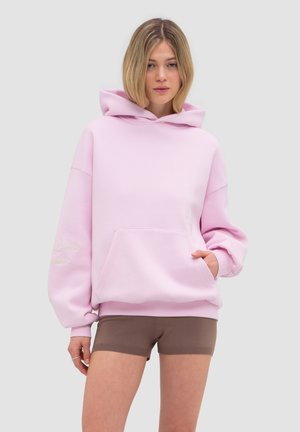 Młoda kobieta z blond bobem nosi oversize pastelowo różową bluzę z kapturem i dopasowane brązowe szorty na jednolitym tle.