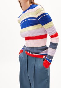 Kvinna med händerna i fickorna iklädd en ribbad flerfärgad randig tröja och lösa blå byxor mot vit bakgrund.