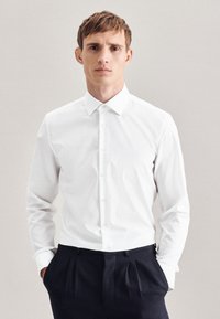 Camicia bianca a maniche lunghe con colletto button-down, caratterizzata da una texture liscia e un taglio aderente, abbinata a pantaloni scuri.