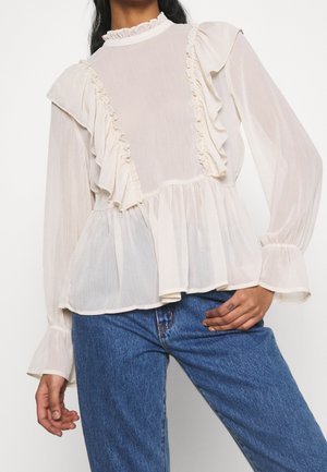 Biała, teksturowana bluzka z falbanami na ramionach i z przodu, z długimi rękawami i zakończona baskinką; w zestawieniu z niebieskimi dżinsami.