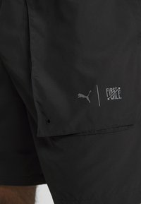 Mustat shortsit, joissa on sileä pinta ja cargo-tasku. Mukana Puma-logo ja "FIRST MILE" -brändäys harmaana sivussa.