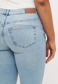 Svijetloplave traperice s glatkom teksturom, koje imaju brendiranu smeđu kožnu etiketu, stražnje džepove i uski kroj.