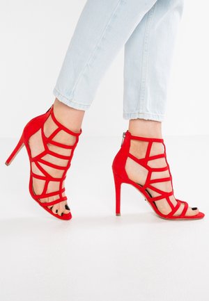 Czerwone zamszowe sandały na wysokim obcasie z misternym, krzyżowym wzorem, otwartym noskiem i zapięciem na zamek z tyłu.