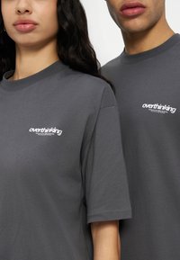 Μερική θέα δύο ατόμων που φορούν σκούρα γκρι μπλουζάκια με τη λέξη «overthinking» τυπωμένη σε λευκό στο στήθος.