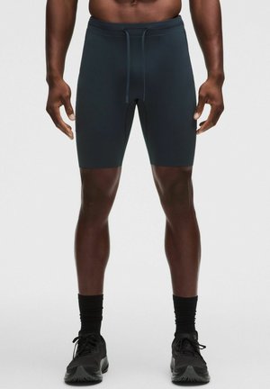 Atletische man draagt donkere nauwsluitende shorts, zwarte sokken en zwarte hardloopschoenen, staand op een effen lichte achtergrond.