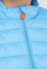 Světlomodrá bunda s péřovým vypěněním má horizontální švy, vysoký límec a kontrastní oranžový zip. Materiál vypadá hladký a lesklý.