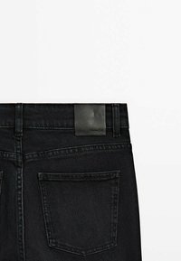 Vue de dos d'un jean en denim noir montrant la poche arrière droite, les passants de ceinture et un petit patch de marque en cuir noir sur la ceinture.