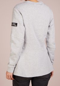Szara bluza z długimi rękawami, zaokrąglonym dołem oraz czarną haftowaną naszywką "KARL" na rękawie. Miękka tekstura, luźny krój.