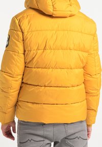 Personne portant une veste matelassée jaune vif avec une capuche et un écusson sur le bras gauche, associée à un pantalon gris clair avec des poches arrière visibles.