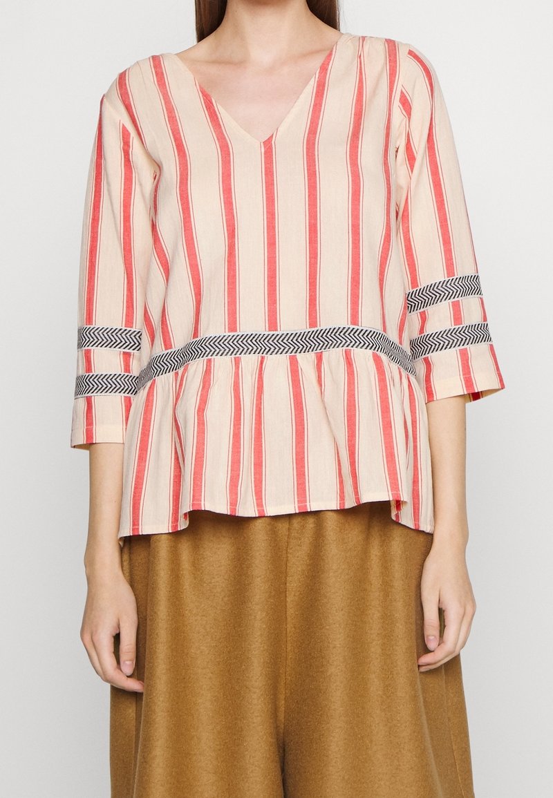 Femme portant un chemisier à rayures verticales beige et rouge avec des bandes à motif chevron à la taille et aux manches, associé à un pantalon large marron.