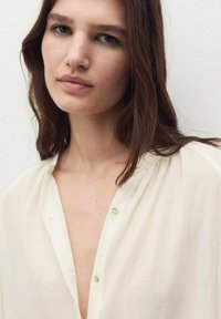 Chemise légère de couleur crème avec un décolleté en V profond, des accents de boutons et une texture subtile. Le tissu semble doux et fluide.