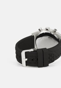 Correa de reloj de goma negra con superficie texturizada y hebilla metálica plateada. Incluye botones de cronógrafo en la parte trasera de la caja de acero inoxidable.