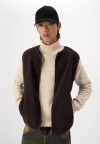 Braunfarbene Fleece-Weste mit Reißverschluss, getragen über einem cremefarbenen, gerippten Rollkragenpullover. Das Model trägt außerdem eine schwarze Mütze. Einfaches Design, ohne Muster.