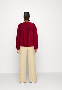 Rød tekstureret bluse med puffede ærmer, løs pasform og en samlet rygdesign, kombineret med brede beige bukser og hvide sneakers.