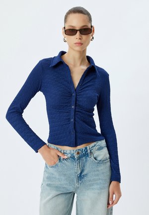 Marineblauwe overhem shirt met een textuur van gekreukt stof. Lange mouwen, getailleerde pasvorm, contrasterende lichte denim jeans.