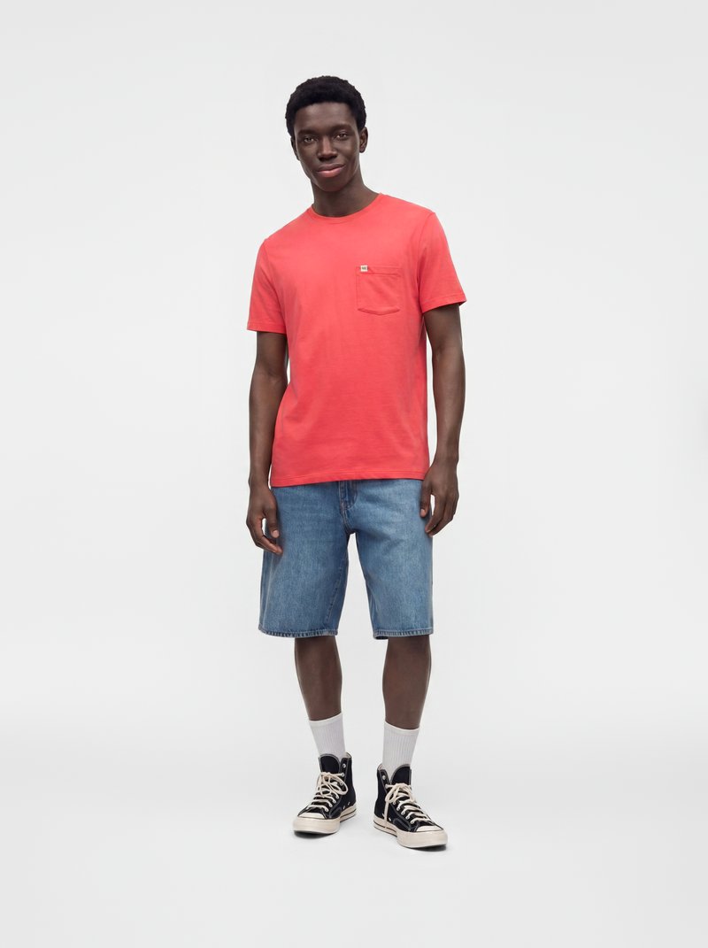 Jeune homme debout, portant un t-shirt rouge, un short en jean bleu, des chaussettes blanches et des baskets montantes noires sur un fond uni clair.