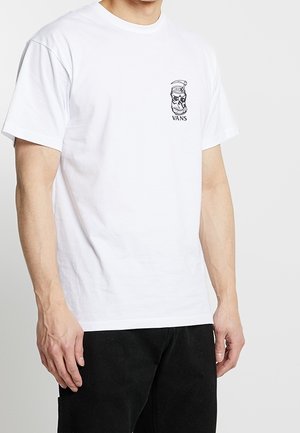 Mężczyzna ubrany w białą koszulkę Vans z małym czarnym logo czaszki w czapce marynarskiej na lewej piersi, w połączeniu z czarnymi spodniami.