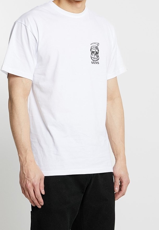 Homme portant un t-shirt blanc Vans avec un petit logo noir de crâne et de chapeau de marin sur le côté gauche de la poitrine, associé à un pantalon noir.